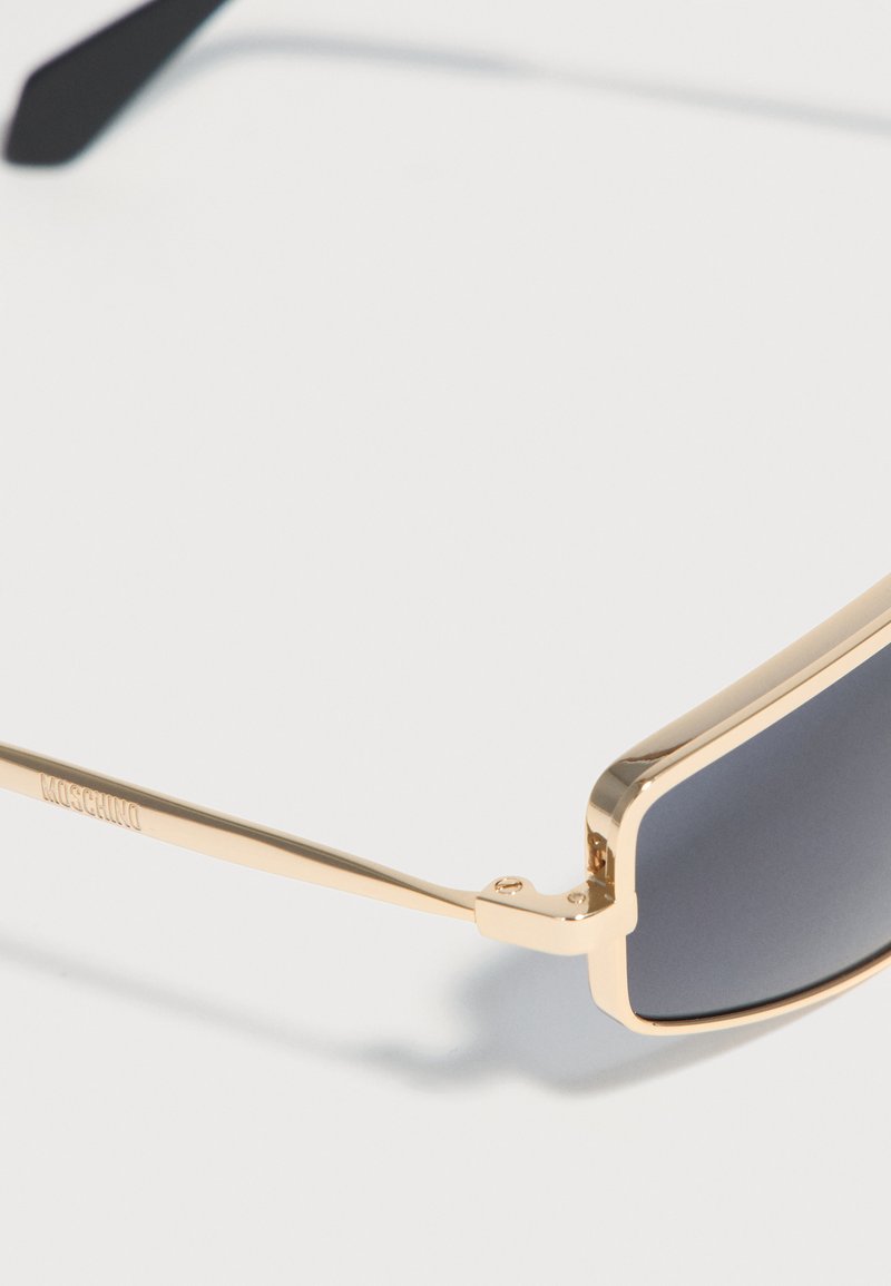 Lunettes de soleil rectangulaires à monture dorée avec verres foncés et branches fines, une branche gravée du nom de la marque Moschino.