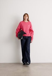 Pink sweatshirt with a crew neck and ribbed cuffs, paired with dark blue wide-leg jeans and black slip-on shoes, holding a black bag.