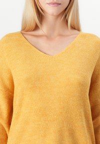 Donna che indossa un maglione giallo a coste con scollo a V, che mostra il collo e la parte superiore del petto, con capelli biondi lisci.