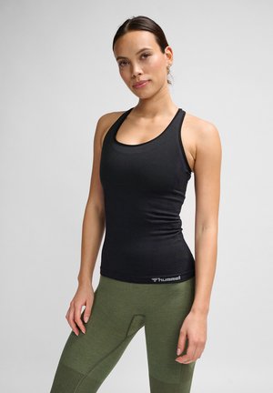 Kobieta stojąca w czarnym topie bez rękawów i zielonych legginsach z fakturą, pozująca na jednolitym jasnoszarym tle.