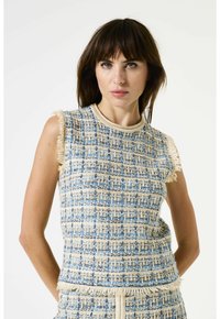 Vrouw met bruin haar, gekleed in een mouwloze blauwe en beige getextureerde tweedtop met franjes, staande tegen een witte achtergrond.
