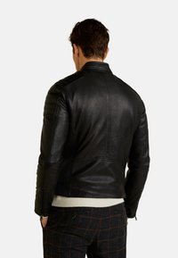 Schwarze Leder-Motorradjacke mit gesteppten Schulterdetails, gerippten Seitenpanele und Stehkragen, getragen über einem weißen Pullover.