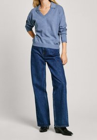 Femme portant un pull à col en V bleu clair, un jean large bleu foncé et des bottines à talons noires, debout avec une main derrière le dos.