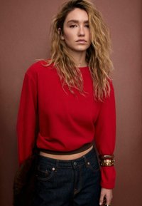 Maglione rosso corto con maniche lunghe e scollo a coste, abbinato a jeans di denim scuro. Accenti marroni evidenti e un braccialetto spesso al polso.