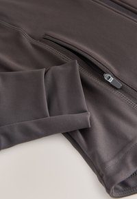 Material sport de culoare gri închis cu mâneci lungi prevăzute cu manșetă și buzunar cu fermoar. Textură fină cu detalii de cusături de-a lungul cusăturilor.