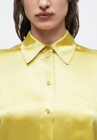 Prim-plan al unei persoane care poartă o cămașă galbenă de satin cu nasturi și guler ascuțit, având un piercing pe buza superioară.