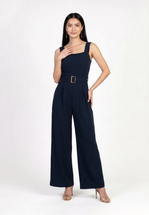 Kvinde står iført en ærmeløs marineblå jumpsuit med vide ben, firkantet udskæring, bælte med spænde og gyldne høje hæle sandaler.