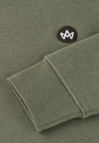 Sweatshirt vert olive avec une manchette côtelée et un patch circulaire marron arborant un logo blanc. Le tissu semble doux avec une texture lisse.