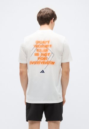 Maglietta bianca in cotone con testo arancione e nero sul retro, elementi di design geometrici e un piccolo logo vicino all'orlo.