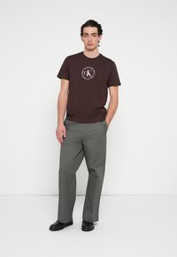 Brązowy bawełniany t-shirt z białym okrągłym logo, zestawiony z szarymi spodniami o szerokich nogawkach. Model stoi na jednolitym tle, ubrany w czarne buty.