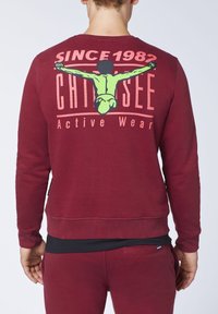 Sudadera marrón con cuello redondo, que presenta un estampado gráfico en la parte posterior. Muestra letras en verde y rosa y una ilustración.