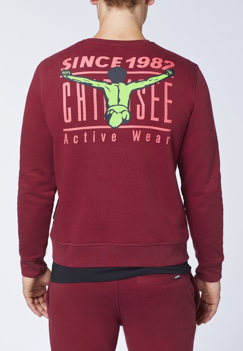 Sudadera marrón con cuello redondo, que presenta un estampado gráfico en la parte posterior. Muestra letras en verde y rosa y una ilustración.