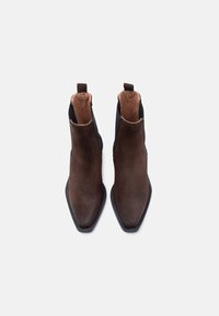 Ζεύγος μποτών αστραγάλου από σκούρο καφέ σουέντ με μυτερά δάχτυλα και μαύρα ελαστικά πλαϊνά πάνελ σε λευκό φόντο.