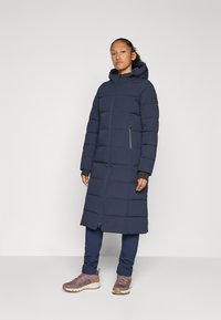 Marineblå lang puffjakke med hette, med horisontale lappepartier, frontglidelås og sidelommer; brukt sammen med marineblå bukser og brune sko.