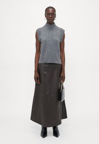 Maglione grigio senza maniche con collo alto, orlo a coste, abbinato a una lunga gonna in pelle marrone. Il modello tiene una borsa nera con dettagli in catena.