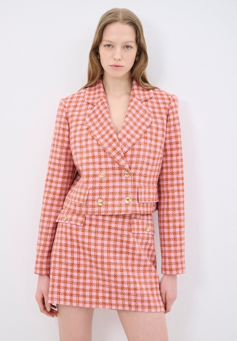 Dwurzędowa marynarka w różowo-pomarańczowy wzór houndstooth z złotymi guzikami, w zestawie z dopasowaną mini spódnicą. Teksturowany materiał, dopasowany krój.