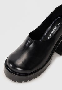 Μαύρο δερμάτινο παπούτσι πλατφόρμα με χοντρό τακούνι και αυλακωτή σόλα, με λευκά ραφτά εσωτερικά και το όνομα της μάρκας τυπωμένο στην εσωτερική σόλα.