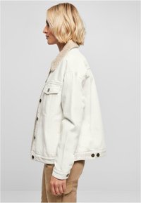 Veste en denim blanche avec un col en fausse fourrure, fermeture à boutons, deux poches avant et coupe décontractée. Le tissu a une texture lisse.