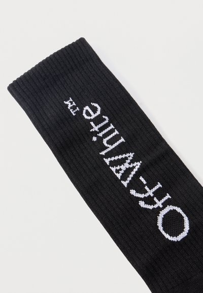 Chaussette noire côtelée avec le logo "Off-White™" tricoté en blanc sur le côté, sur un fond blanc uni.