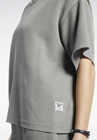 Šedé ribované tričko s krátkým rukávem a volným střihem. Obsahuje malou štítkovou etiketu Reebok na spodním lemu a má hladkou texturu.