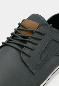 Scarpa sintetica grigia con superficie testurizzata, accento in pelle marrone e design con lacci scuri, caratterizzata da un profilo basso e suola in gomma bianca.