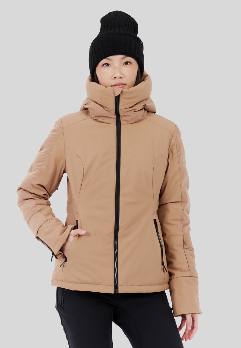 Chaqueta aislante marrón con un cuello alto, cremallera negra y dos bolsillos laterales. Combinada con un gorro de punto negro.