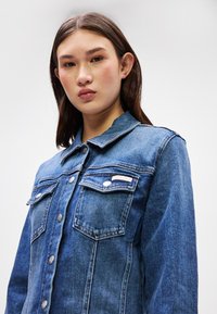 Τζάκετ από τζιν σε μεσαίο μπλε χρώμα με γιακά, δύο τσέπες στο στήθος, κούμπωμα με ασημένια κουμπιά και ετικέτα μάρκας στην αριστερή τσέπη.