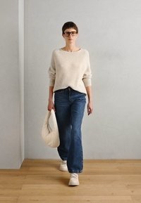 Suéter de punto acanalado color crema, jeans de pierna ancha azules, zapatillas blancas, sosteniendo un bolso hobo beige, de pie sobre un suelo de madera contra una pared clara.