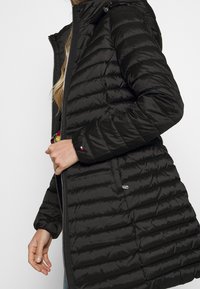Schwarze gepolsterte Jacke mit Kapuze, die ein elegantes Design, ein strukturiertes Steppmuster und silberne Beschläge an dem vorderen Reißverschluss und den Taschen aufweist.