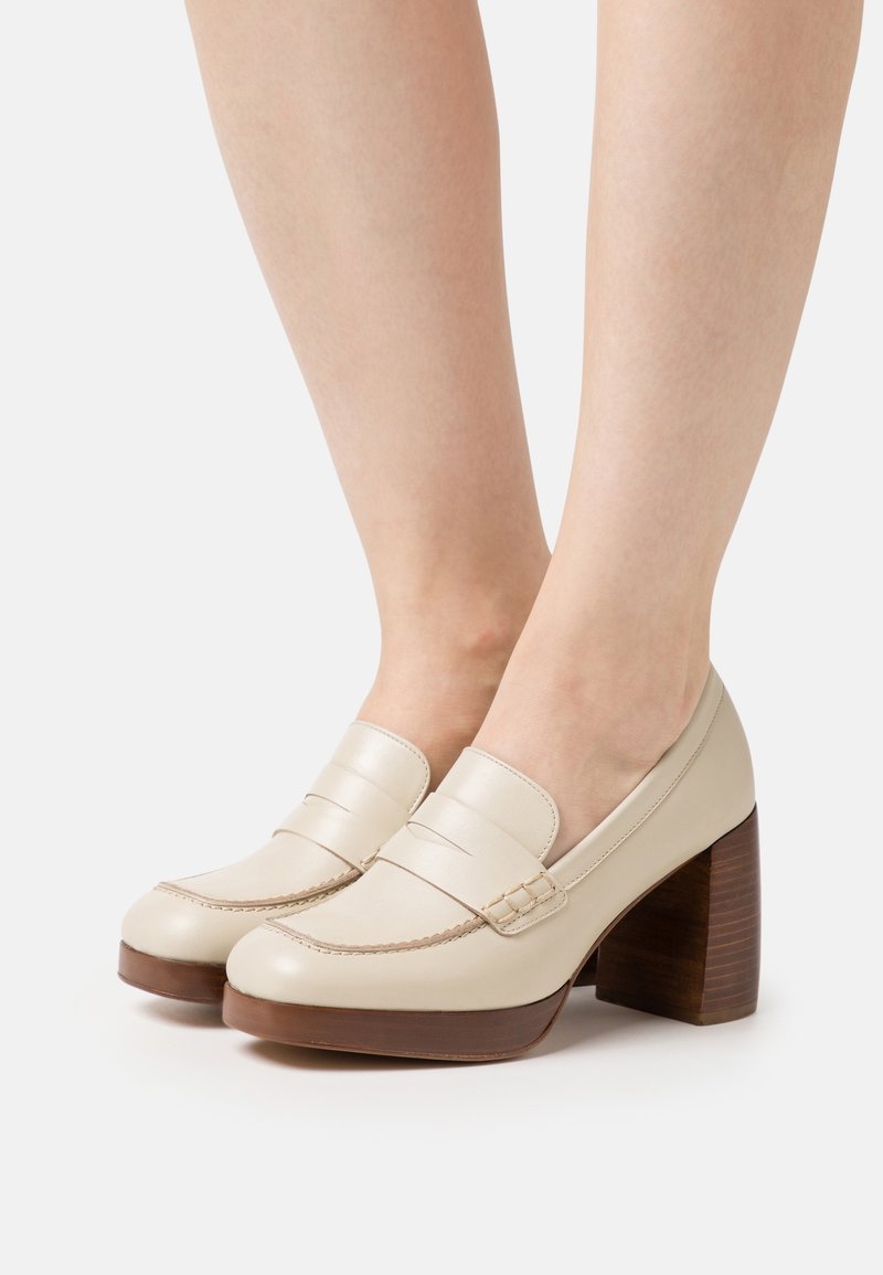 Mocassins à plateforme en cuir beige avec talons en bois épais portés pieds nus, présentés sur un fond blanc uni.