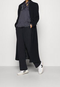 Cappotto lungo nero sopra una blusa blu navy a pois con cravatta a fiocco, abbinato a pantaloni neri larghi e sneakers bianche con dettagli neri.