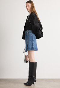 Schwarze Bomberjacke über einem schwarzen Oberteil, kombiniert mit einem blauen Jeansrock. Das Model trägt hohe schwarze Stiefel mit Absatz und hält eine metallische Handtasche.
