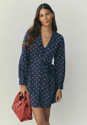 Rochie bleumarin cu buline, mâneci lungi, închidere cu nasturi în față și talie cu cordon. Asortată cu o geantă roșie cu accesorii aurii.