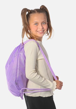 Lächelndes Mädchen mit blauen Augen und Zöpfen, trägt ein cremefarbenes langärmeliges Oberteil und schwarze Hosen, trägt einen violetten Kordelzug-Rucksack.