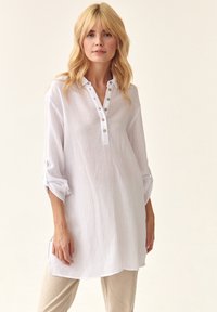 Hvid, struktureret tunika skjorte med knaplukning, oprullede ærmer og sidesplitter, båret over lyse beige bukser.