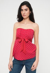 Κόκκινο τοπ χωρίς τιράντες με λευκές πουά, πιετάσα στο μπούστο και φαρδιά φιόγκο στη μέση. Συνδυασμένο με μπλε τζιν παντελόνι.