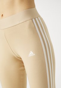Beiga leggings med en bred midjeband, tre vita ränder längs sidorna och en vit Adidas-logotyp på underbenet.