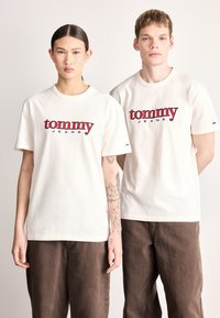 Białe bawełniane koszulki z logo "tommy JEANS" w czerwonym i czarnym kolorze, z krótkimi rękawami i luźnym krojem. Noszone z brązowymi spodniami.