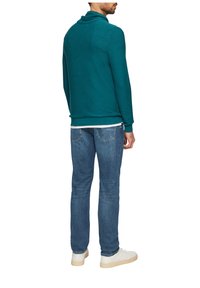 Felpa turchese con collo alto, abbinata a jeans azzurri e sneakers bianche. Il tessuto appare testurizzato e lavorato a maglia.