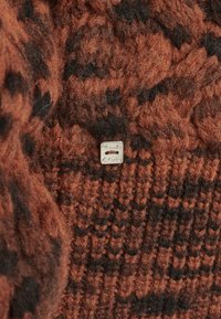 Tessuto a maglia di un maglione in tonalità marrone e nera, caratterizzato da un motivo testurizzato e un piccolo tag d'argento con incisione.