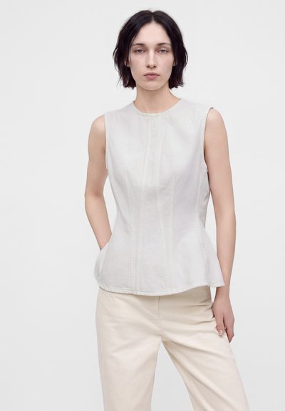 Jeune femme aux cheveux courts et foncés portant un haut blanc ajusté sans manches et un pantalon beige clair, debout devant un fond uni.
