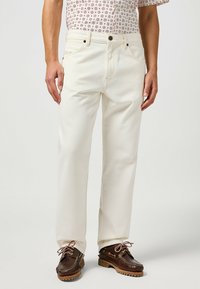 Vaqueros de mezclilla blanca con corte recto, cinco bolsillos y herrajes plateados. Llevados con zapatos marrones con cordones decorativos y una suela texturizada.