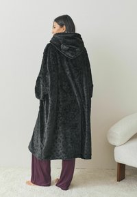 Oversized szlafrok z czarnym nadrukiem w panterkę, wykonany z pluszowego materiału, charakteryzujący się miękką teksturą i luźnym krojem, w połączeniu z luźnymi fioletowymi spodniami.