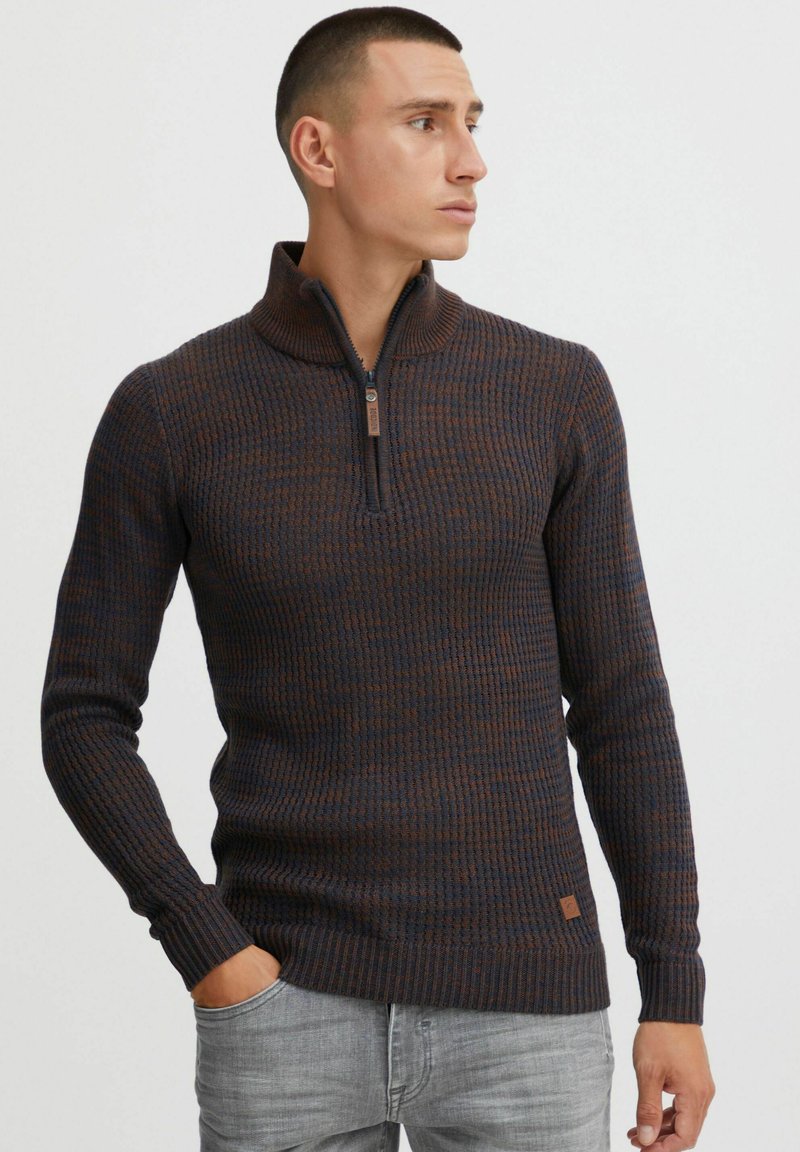 Strickpullover mit einem halben Reißverschluss am Kragen, der ein strukturiertes Muster in dunklem Blau und braunen Farbtönen aufweist. Enthält ein kleines Markenakzent am Saum.