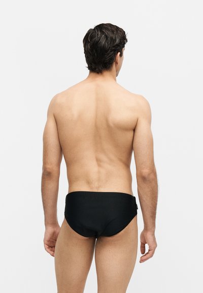 Άνδρας με κοντά σκούρα μαλλιά που στέκεται με την πλάτη, φοράει μαύρο μαγιό σε απλό ανοιχτό γκρι φόντο.