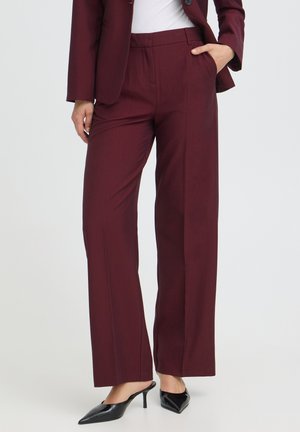 Femme portant un pantalon de costume bordeaux avec blazer assorti, un haut blanc et des chaussures à talons noires, debout avec une main dans la poche.