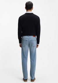 Čierny sveter s dlhým rukávom, svetlomodré nohavice s rovnými nohavicami, hnedý štítok na opasku nohavíc a hnedé topánky. Zobrazený z pohľadu zozadu.