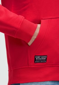 Sudadera roja con puños y dobladillo acanalados, que presenta un bolsillo frontal tipo canguro y una etiqueta negra con texto blanco en el lateral.