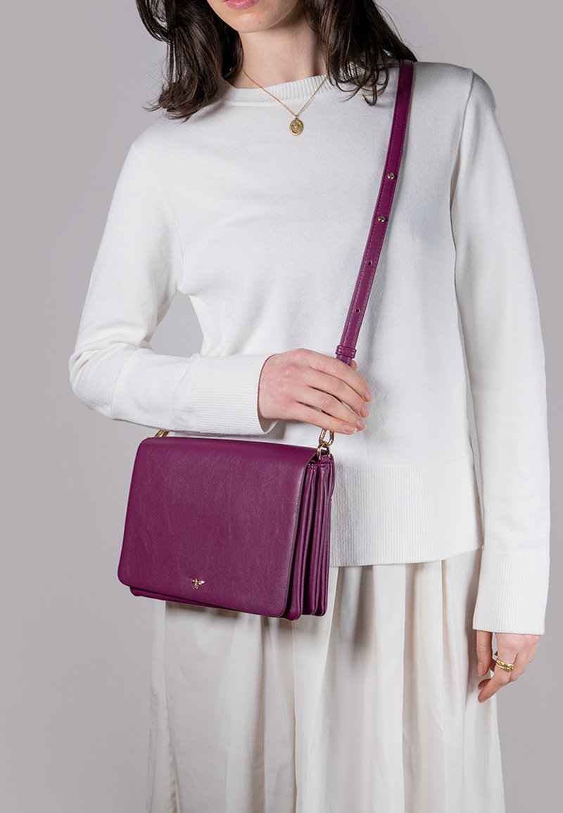 Lila läder crossbody-väska med en strukturerad design, guld detaljer och justerbar rem, ihop med en vit stickad tröja och en krämfärgad kjol.