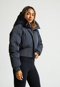 Giacca puffer nera corta con colletto alto, chiusura con zip e tasche laterali. Presenta un accento testurizzato all'orlo e ai polsini.